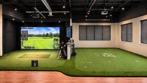 419 Golf Manufacturing's indoor golf testing facility Trackman 4 and a PuttView putting simulator green, a large projection screen showing a golf course, and a golf bag on a stand. The room has black walls, windows with dark blinds, and a wood floor.