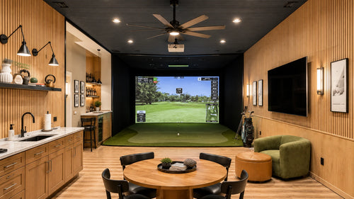 A luxury home golf simulator room with a large projection screen showing a golf course, a green turf hitting surface, a round wooden dining table with black chairs, a green armchair, and a built-in wet bar on the left.
