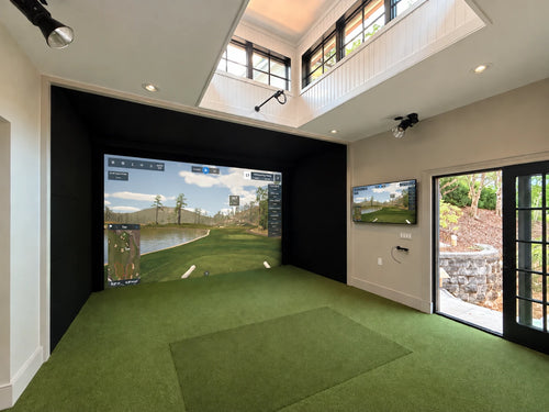An two level residential golf simulator room with a 2nd floor balcony, green turf hitting surface, a large black-framed projection screen showing a golf course, a smaller wall-mounted screen, and glass doors looking out to a landscaped outdoor area.