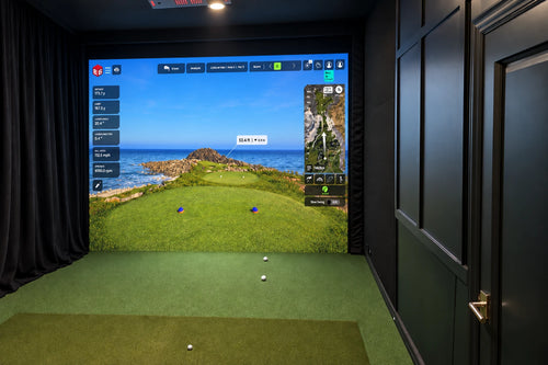 A custom indoor golf simulator room with a large projection screen with a golf course image that goes all the way to the floor, green turf hitting surface, and black paneled wood trim with curtains on the left.