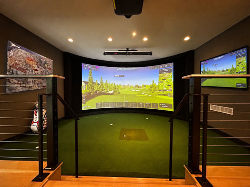 A step down basement home golf simulator setup with a curved projection screen showing a golf course, a green turf hitting surface, side monitors, and dark wood walls.