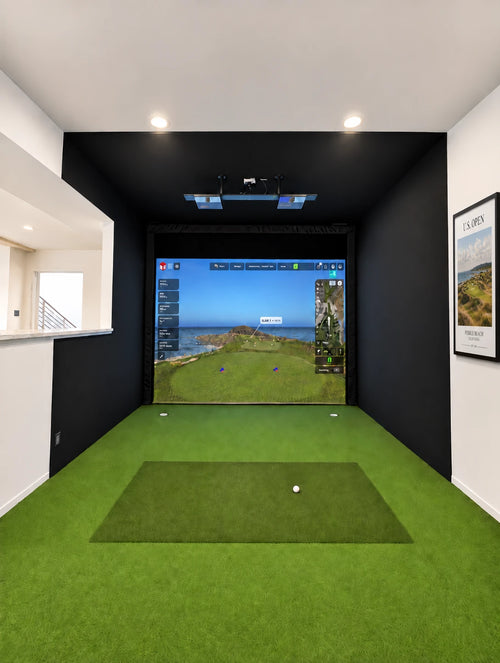 An How much room do you need for an indoor golf simulator? This 14' wide x 17' deep sim room features green turf, black padded walls, a screen showing a golf course simulation, and a golf ball on the hitting mat with a Framed US Open golf print on the side wall.