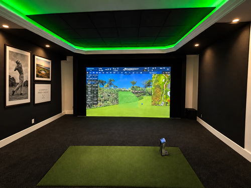 A Foresight GCuad golf simulator room with a large projection screen showing a golf course view, featuring a green stance mat on a black putting turf, and green LED lighting in the ceiling.