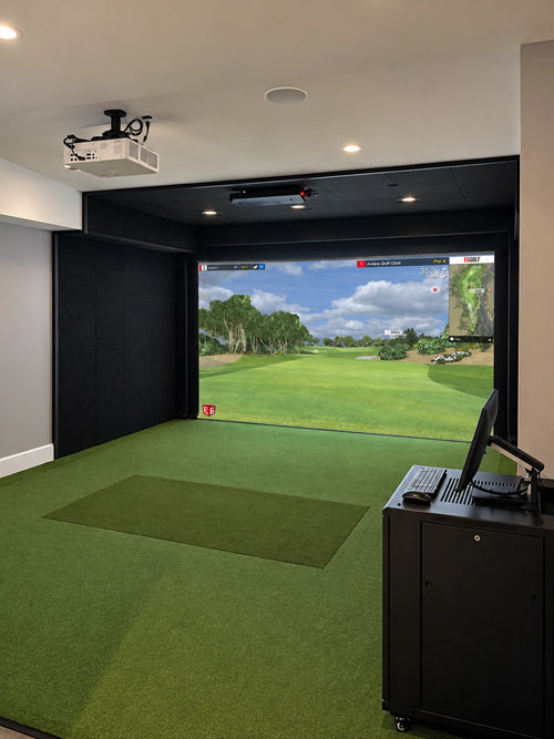 Custom Uneekor Eye XO2 indoor golf simulator room with a green turf hitting mat, black enclosure walls, a ceiling-mounted projector, and a computer cart with a monitor in the foreground. A large simulated golf course screen is visible behind the room.