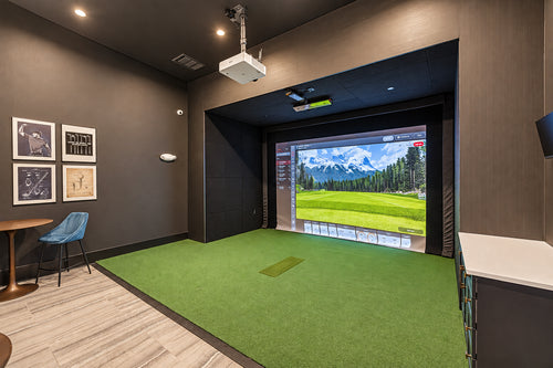 A luxury apartment building with a Uneekor Eye XO2 golf simulator room with a green turf hitting area, dark wood walls, framed wall art, a small table and chair, and a large projection screen showing a golf course landscape.