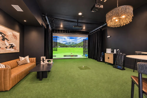 A luxury apartment complex Lawrence Wallen Park's golf simulator setup featuring a large projection screen showing a golf course, artificial turf flooring, a brown leather sofa, and golf clubs on a stand.