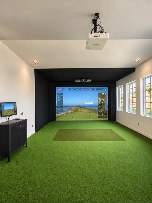 A commercial indoor golf simulator room at the Dexter Clubhouse with green turf flooring, acoustic tiles, a large projection screen showing a golf course simulation, a ceiling-mounted projector, and side windows.