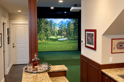 A residential custom Foresight Falcon golf simulator featuring a large projected golf course scene, with a green carpeted floor, granite countertops, framed wall art, and a whiskey bottle and glasses on a tray in the foreground.