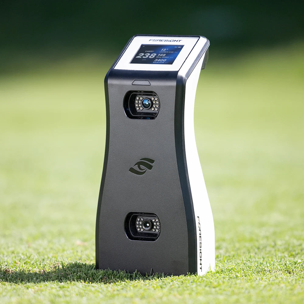 A Foresight GC2 monitor standing upright on a green grass hitting tee, with its digital screen illuminated and two front-facing cameras visible.
