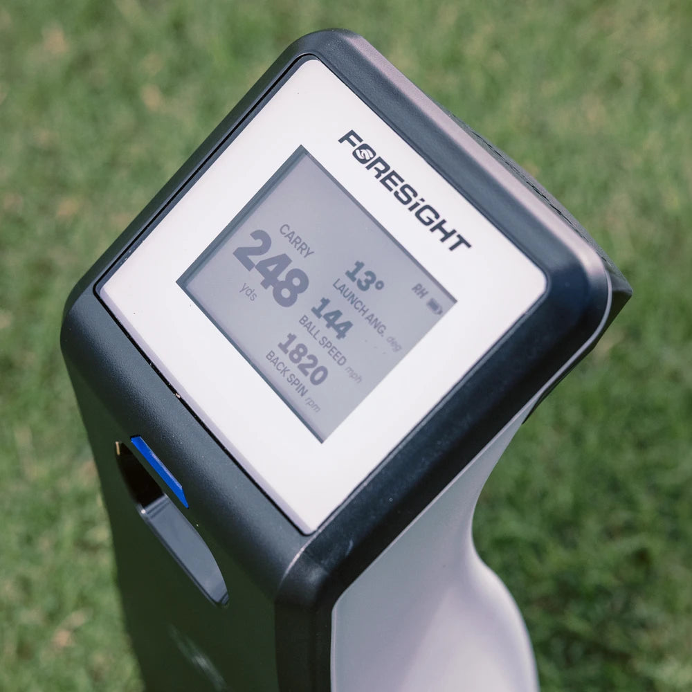 A Foresight golf GC2 launch monitor standing outdoors on grass, showing a close up of the digital screen with carry, launch angle, ball speed, and back spin RPM.