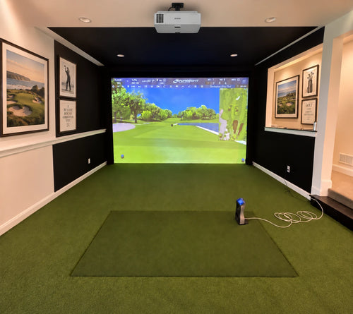 A Foresight GCQuad, residential golf simulator setup in a home office, featuring a green turf hitting mat, a golf ball on a launch monitor, a projector mounted on a black ceiling, and a large screen displaying a golf course simulation.