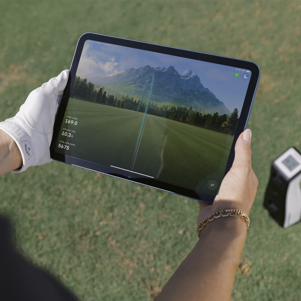 A female golfer wearing a white golf glove holds a tablet displaying the Foresight Premier driving range showing golf trajectory with the Foresight GC2 visible in on the grass in the background.