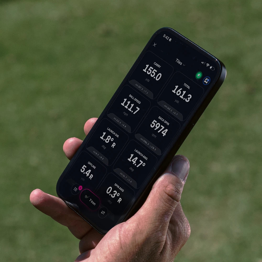 A close-up of a hand holding a smartphone outdoors on a grassy field, displaying the Foresight Premier Tile app with metrics like carry, total, club speed, ball speed, launch direction, launch angle, spin axis, and offline on the screen.