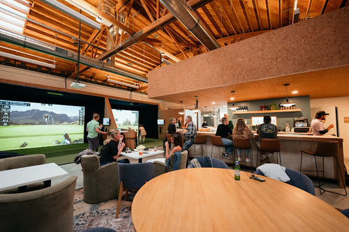 Four Wood Golf in California featuring a modern indoor golf lounge with Uneekor Eye XO2 monitors, large projection screens showing a golf course, people watching and socializing, a bar area with stools, and a high wood ceiling.