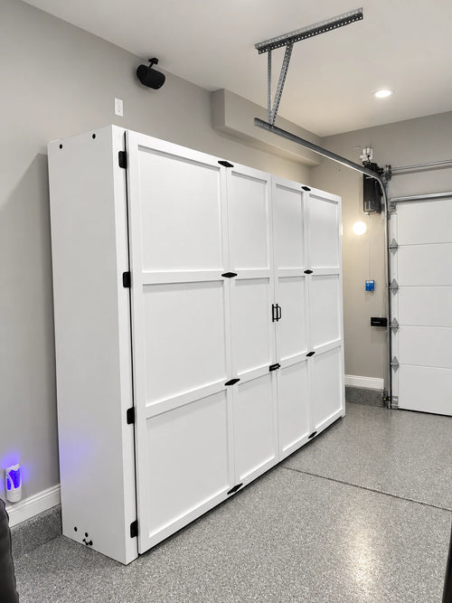 A tall white Murphy retractable golf simulator storage cabinet with multiple door panels and black handles, installed in a garage with gray walls, a gray floor, and a garage door opener system visible.