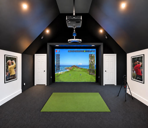 Vaulted ceiling ProTee VX custom golf simulator with a screen showing a virtual golf course, under a projector mounted on the ceiling. A green hitting mat is placed on black turf flooring, with framed golf portraits of Tiger Woods and Jack Nicklaus on the white walls.