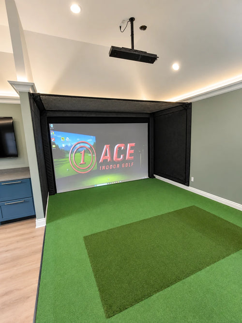 An indoor golf simulator with a top lid/net setup in a home with vaulted ceilings. Features a green turf hitting surface, a large projection screen displaying the 'ACE Indoor Golf' logo, black acoustic wall panels, and a Uneekor Eye XO2 launch monitor. 
