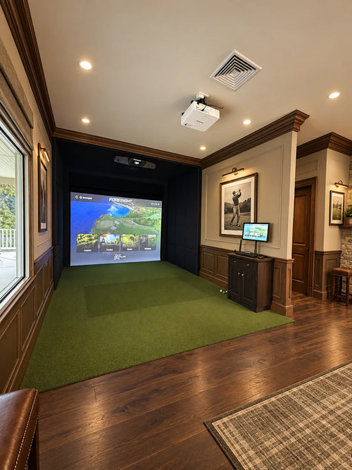 A golf course clubhouse simulator with a large projected golf course scene on a green turf floor, wood-paneled walls, hardwood flooring, a projector mounted on the ceiling, and a small monitor on a console.