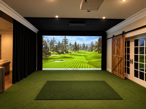 A Uneekor Eye XO@ indoor golf simulator room with a large projection screen showing a golf course, green turf flooring, a black hitting mat, black curtains on the right protecting the family room, and wooden barn-style door on the right protecting glass doors.