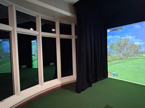 A custom designed indoor golf simulator room with a black theater curtains protecting large glass windows, green turf floor, and a projected golf course scene on a screen.