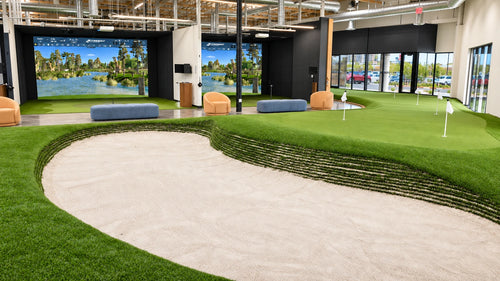 Gretsch Golf Academy two indoor golf simulators behind a practice sand bunker and green in the foreground, artificial green turf, large wall-mounted sim screens, seating, and glass entry doors in the background.