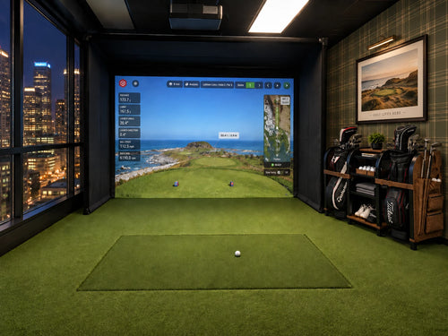 A Uneekor Eye XO2 indoor golf simulator room with a green turf hitting mat, a large back-of-house screen showing a golf course simulation, golf bags on a rack, and city windows at night in the background.