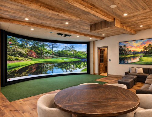 Luxury home golf simulator room and home theater with green turf, wood ceilings, sectional couch, table and chairs, and golf artwork on the wall.