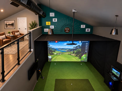 An indoor golf simulator setup in a loft-style room, featuring a large projection screen showing a golf course, a green hitting mat, black sweep curtains, and a modern living area visible on the upper level.