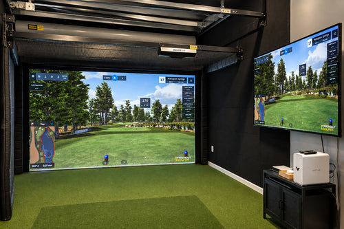The Lake Presidential indoor-outdoor golf simulator hitting bay with retractable doors and golf simulator screen. The large projection screen is showing a golf course. There is also a smaller side flat screen TV, a green turf floor, and a white Vitus computer on a black stand.