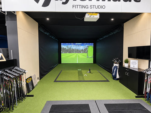 PGA Tour Superstore's indoor golf fitting studio with a green turf hitting mat, a large projection screen showing a golf course, golf clubs displayed on racks, and a fitting studio setup with a sign reading 'TaylorMade FITTING STUDIO'.