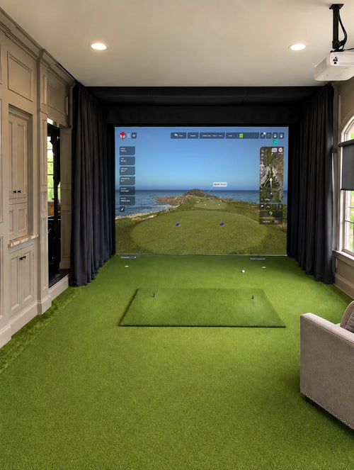 A Trackman IO custom home golf simulator setup in a wood paneled office, featuring a green turf hitting mat, dark curtains, a large projection screen showing a golf course, and a gray armchair in the foreground.
