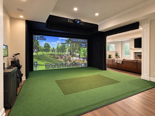 Foresight Falcon home golf simulator setup in a modern dinning room, featuring a large projection screen showing a golf course, black curtains, a green turf hitting mat, and a large opening to the family room.
