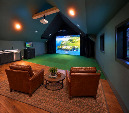 A residential golf simulator setup in a dark room with two brown leather armchairs facing a large projection screen. The room has wood floors, a patterned area rug, a green turf, and a black acoustic tile ceiling with a projector and launch monitor mounted above.