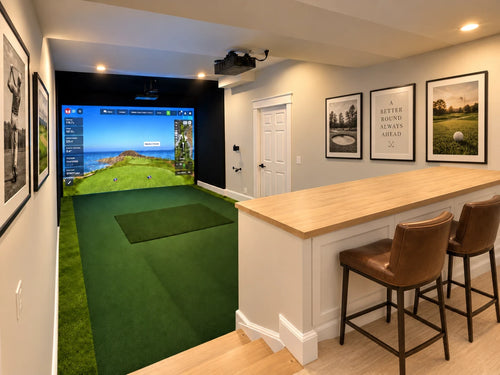 A step down, dug-out,  basement  golf simulator featuring a large indoor golf simulator screen showing a golf course, positioned next to a white bar counter with two brown leather bar stools. The room has light gray walls adorned with framed pictures, and the floor is light wood.