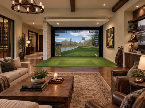 A luxury home golf simulator with a stone side wall and large projection screen displaying a golf course, with a green turf hitting mat and golf ball in the foreground. The room is furnished with leather armchairs, a wooden coffee table, and a patterned area rug.