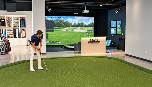 A man at Studio Golf El Segundo in a navy polo shirt and white pants is putting on a PuttView putting green with an indoor golf simulator with a large virtual golf course screen behind him.