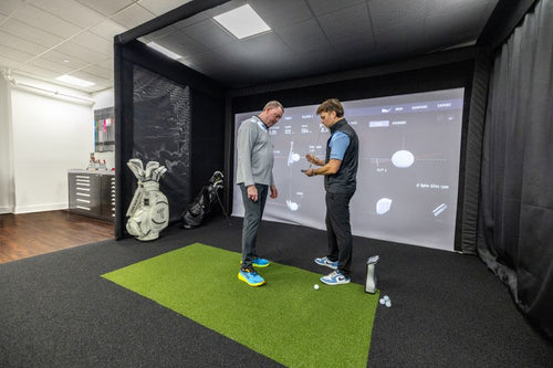 TruSpec Chelsea Pier New York golf simulator club fitting pay. Two men standing on a green turf hitting mat inside a simulator, with one holding a golf ball and the other observing, in front of a large projection screen.