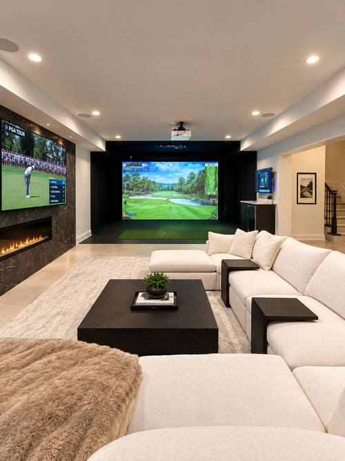 A Uneekor Eye XO2 golf simulator next to a living room setup with a large white sectional sofa, black coffee table, fireplace wall, and a projector screen displaying a golf game. Multiple TVs are mounted on the walls, and the room is lit by recessed ceiling lights.