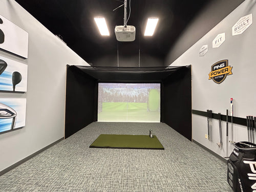 A Worldwide Golf indoor golf simulator room with a black next canopy and enclosure, gray carpeted floor, green turf mat, projector screen showing a golf course, and golf clubs hanging on the side walls.